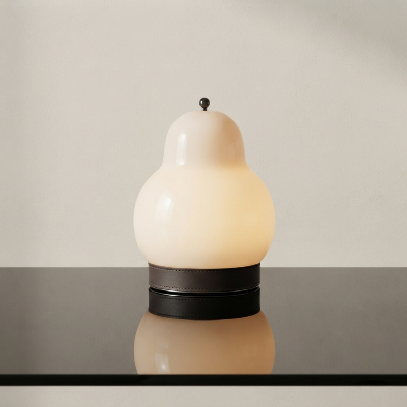 A sculptural table lamp with a pear-shaped frosted glass shade in warm off-white, resting on a dark circular base with a dark leather band, set against a neutral beige background.