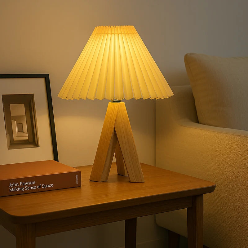 Table lamp with pleated yellow shade on a wooden table, next to a book and framed picture, in a cozy room setting.