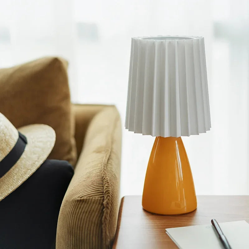Table lamp with pleated white shade on a wooden surface next to a sofa and hat