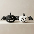 Two plush Halloween pumpkin toys with bat wings, one black and one white, displayed side by side on a light beige surface against a matching neutral background.