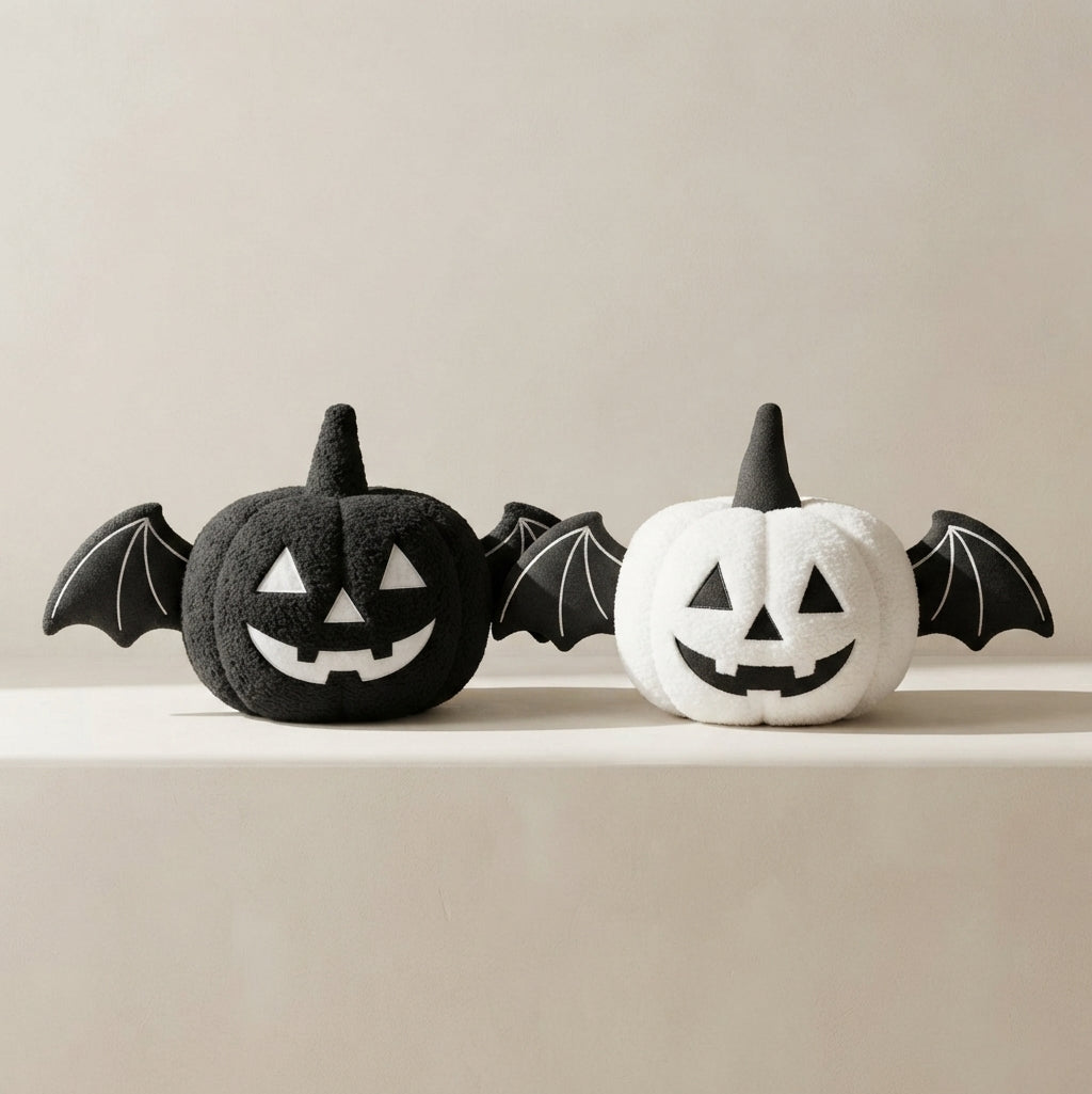 Two plush Halloween pumpkin toys with bat wings, one black and one white, displayed side by side on a light beige surface against a matching neutral background.