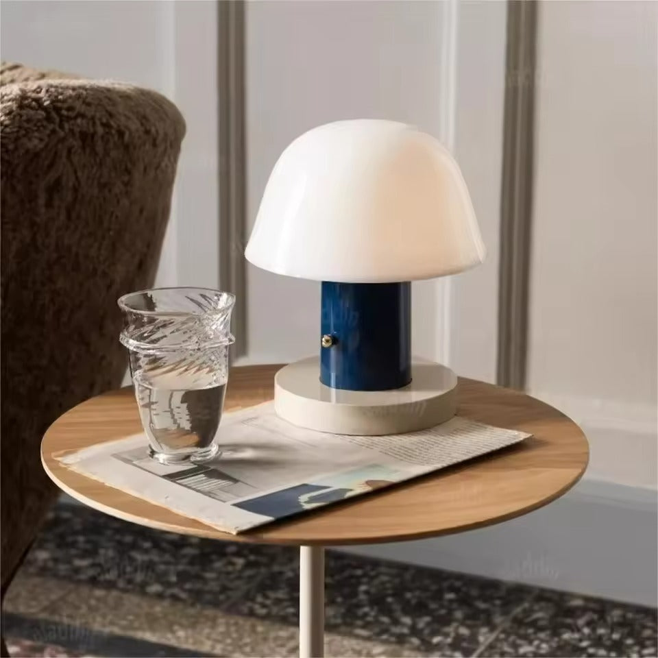 A mushroom-shaped table lamp with a white dome shade and a dark blue cylindrical base, styled on a round wooden side table next to a glass of water in a home interior setting.