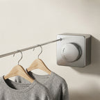 A wall-mounted silver clothes rail with a round control dial, shown holding two gray sweaters on wooden hangers against a plain beige wall.