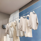 A wall-mounted clothing rail holding several light neutral knit tops and dresses in a styled interior with a blue accent wall, white curtain, and a small lamp.