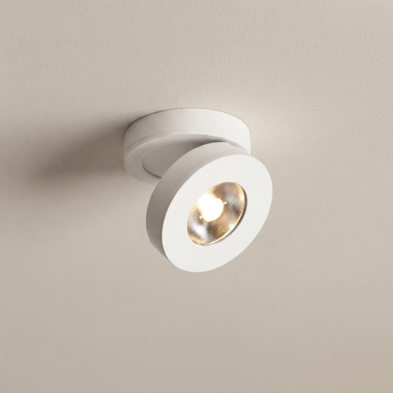 A modern white ceiling spotlight with a circular design, shown mounted on a textured beige wall with the light turned on.
