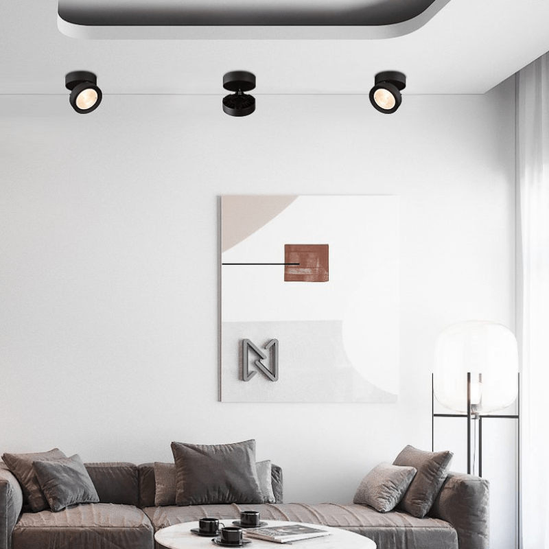 A modern living room scene with a gray sectional sofa, a round coffee table, a black floor lamp, and a large abstract wall art piece hanging above the sofa. The wall art features a white background with soft beige and gray geometric shapes, a small brown square, and a metallic silver stylized letter 'N'.
