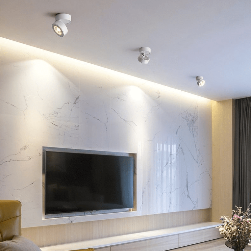 Three white ceiling track spotlight lights mounted on a white ceiling above a modern TV wall with a white marble pattern and warm backlighting.