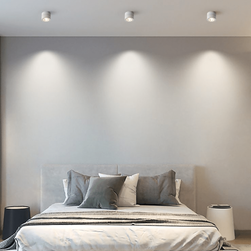 A modern bedroom interior with a neatly made bed, gray and white bedding, a light upholstered headboard, recessed ceiling lights, and two side tables against a plain light gray wall.