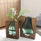 Two green glass vases in wooden holders on a table with a plant.