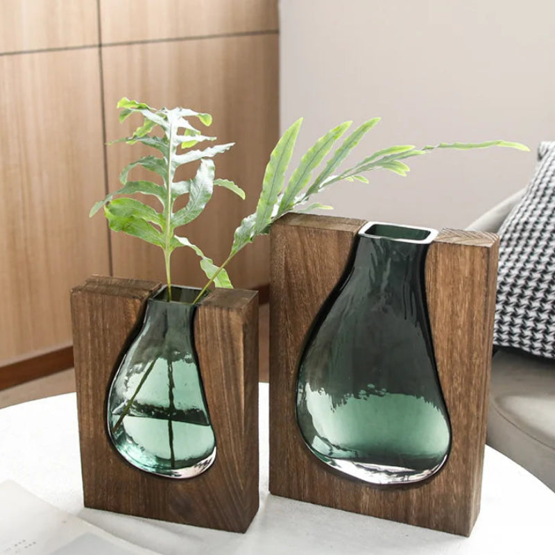 Two green glass vases in wooden holders on a table with a plant.