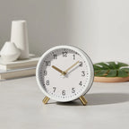 White clock with gold hands on a light surface, with decorative items in the background.