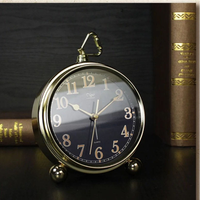 A round vintage-style alarm clock with a gold-toned metal frame and a dark navy blue dial, styled on a dark surface between two antique books.