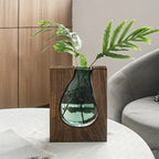 Glass vase with wooden base holding green leaves on a white surface with a neutral background