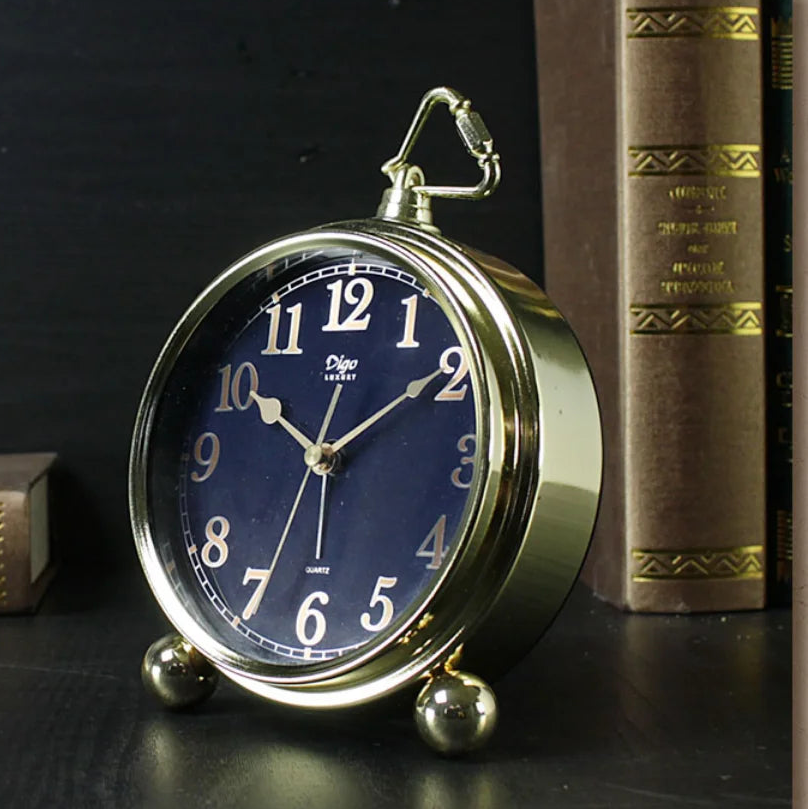 A round vintage-style desk clock with a polished gold-toned metal body, a dark blue clock face, and large white numerals, styled on a dark surface next to old books.