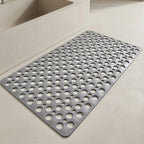 A gray rectangular anti-slip bath mat with a grid of circular drainage holes, shown on a light beige bathroom floor next to a white bathtub.