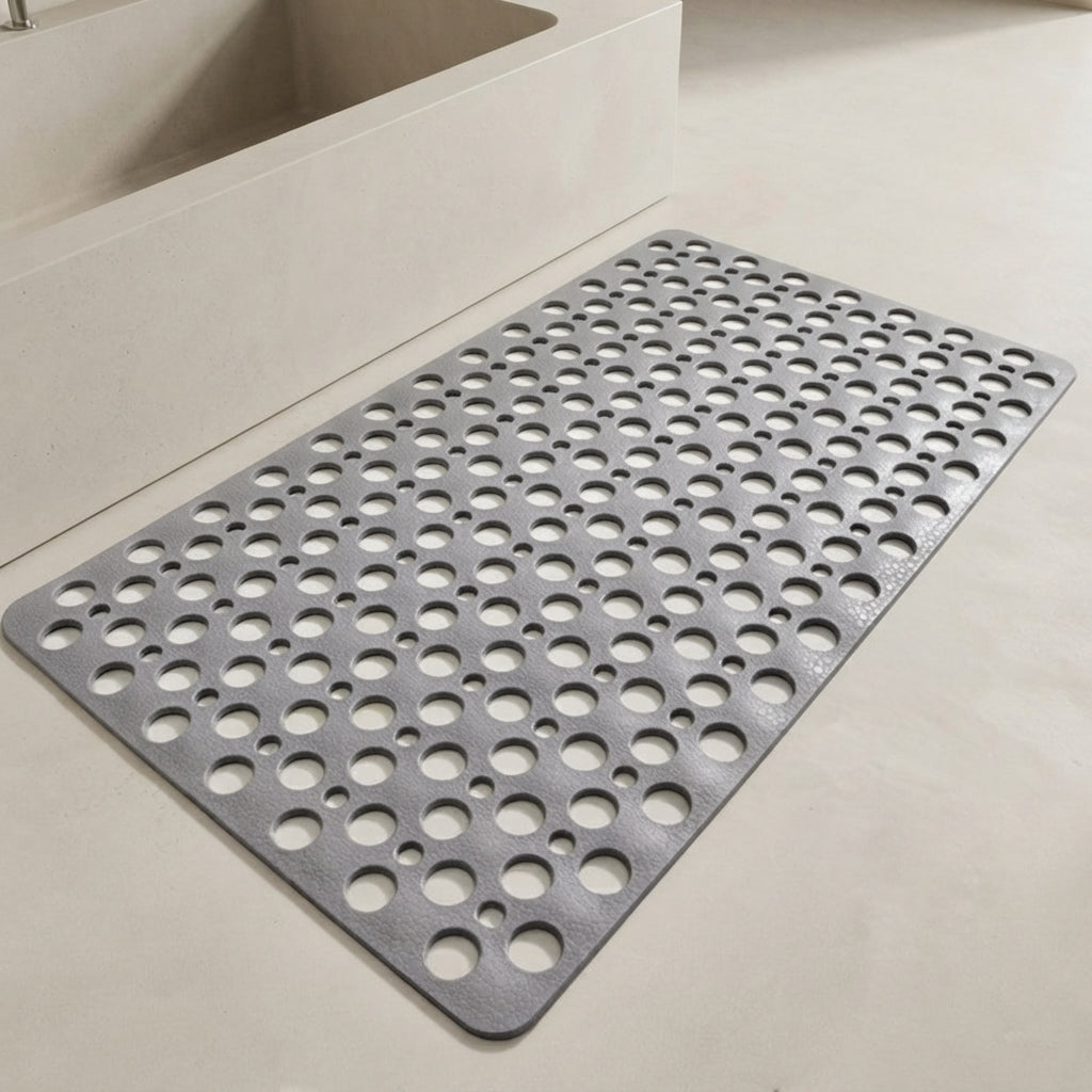 A gray rectangular anti-slip bath mat with a grid of circular drainage holes, shown on a light beige bathroom floor next to a white bathtub.
