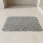 A rectangular gray bath mat with a high-pile looped texture, shown on a light beige floor in a minimalist bathroom setting.