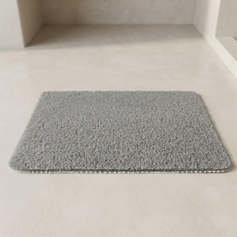 A rectangular gray bath mat with a high-pile looped texture, shown on a light beige floor in a minimalist bathroom setting.