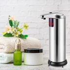 Silver soap dispenser next to a green bottle and white jar on a light wooden surface with a white brick wall background.