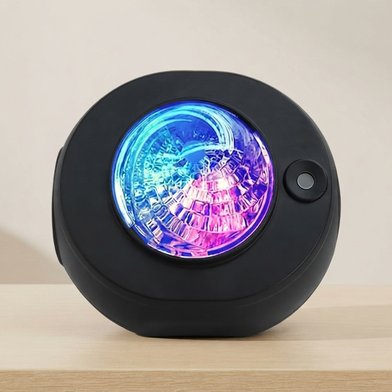 A round black electronic device, likely a portable speaker or light, resting on a light wood surface against a beige background. The front features a circular, illuminated crystal-like lens displaying vibrant blue and purple light patterns, with a small button on the right side.