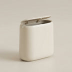 A minimalist, rounded rectangular trash can with a hinged lid in a warm off-white color, set against a matching light beige background.