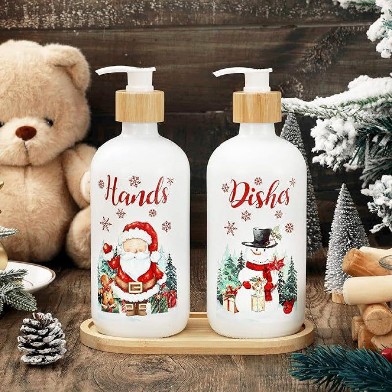 Two white holiday-themed pump bottles on a wooden tray, labeled 'Hands' with a Santa design and 'Dishes' with a snowman design, styled with a teddy bear and winter decorations on a rustic wooden surface.