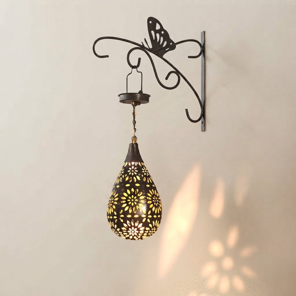 A decorative wall-mounted sconce with a black metal butterfly-shaped bracket, chain, and teardrop-shaped shade. The shade has intricate floral cutout patterns that cast warm light onto the beige wall, creating a shadow pattern.