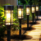 Five black solar-powered outdoor landscape lights with warm yellow LED illumination, arranged along a stone pathway in a garden setting with green foliage and pebbles.