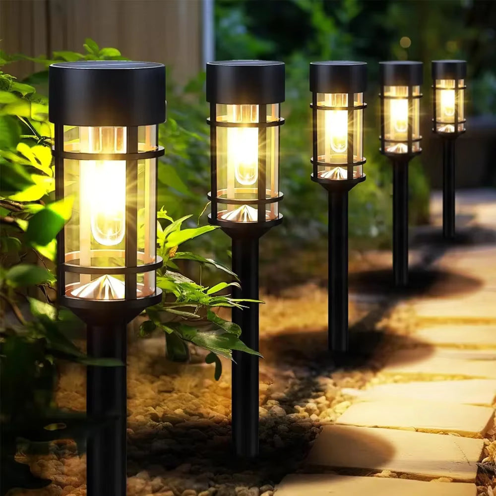 Five black solar-powered outdoor landscape lights with warm yellow LED illumination, arranged along a stone pathway in a garden setting with green foliage and pebbles.