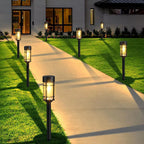 Outdoor solar pathway lights installed along a concrete walkway between a green lawn and a modern house at dusk.