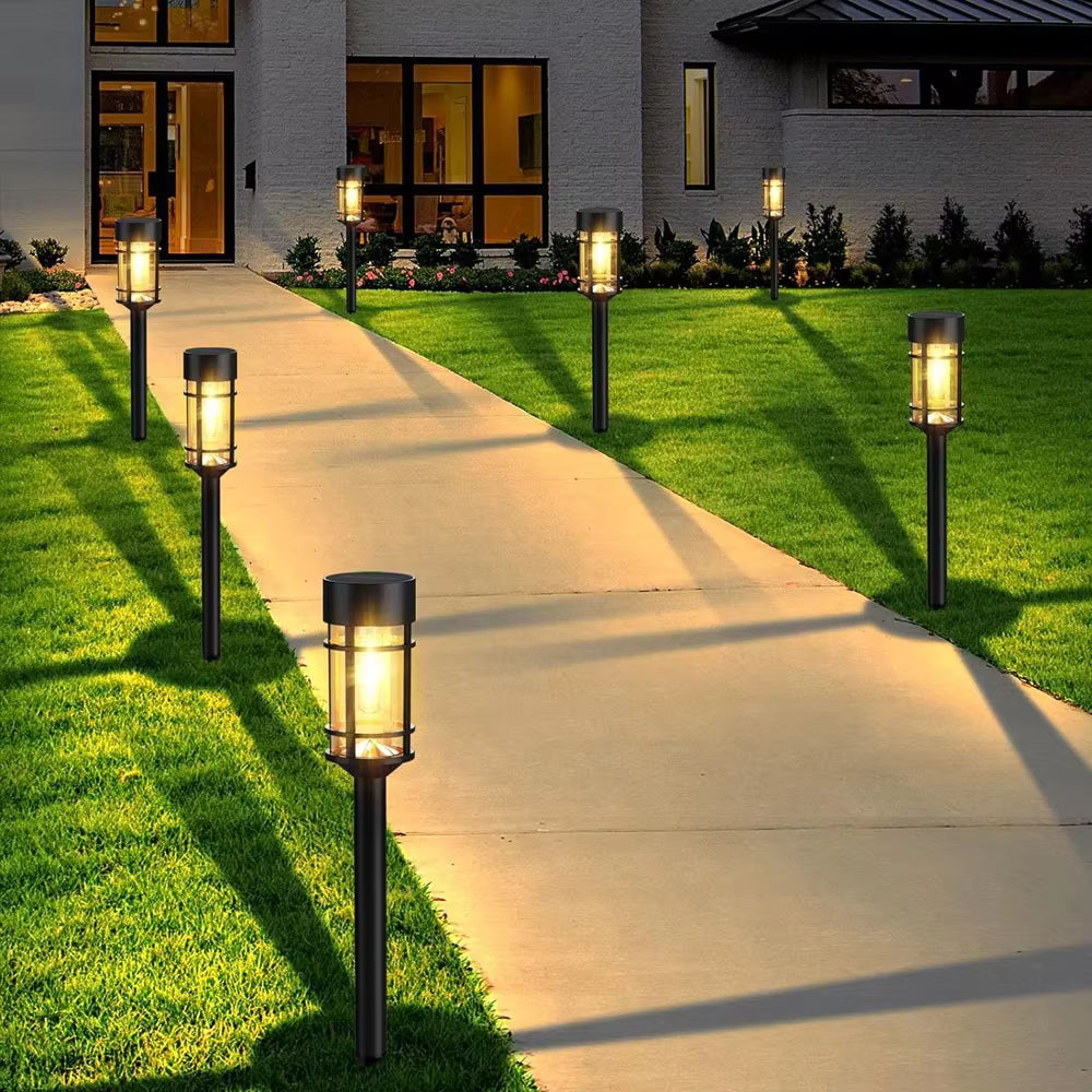 Outdoor solar pathway lights installed along a concrete walkway between a green lawn and a modern house at dusk.