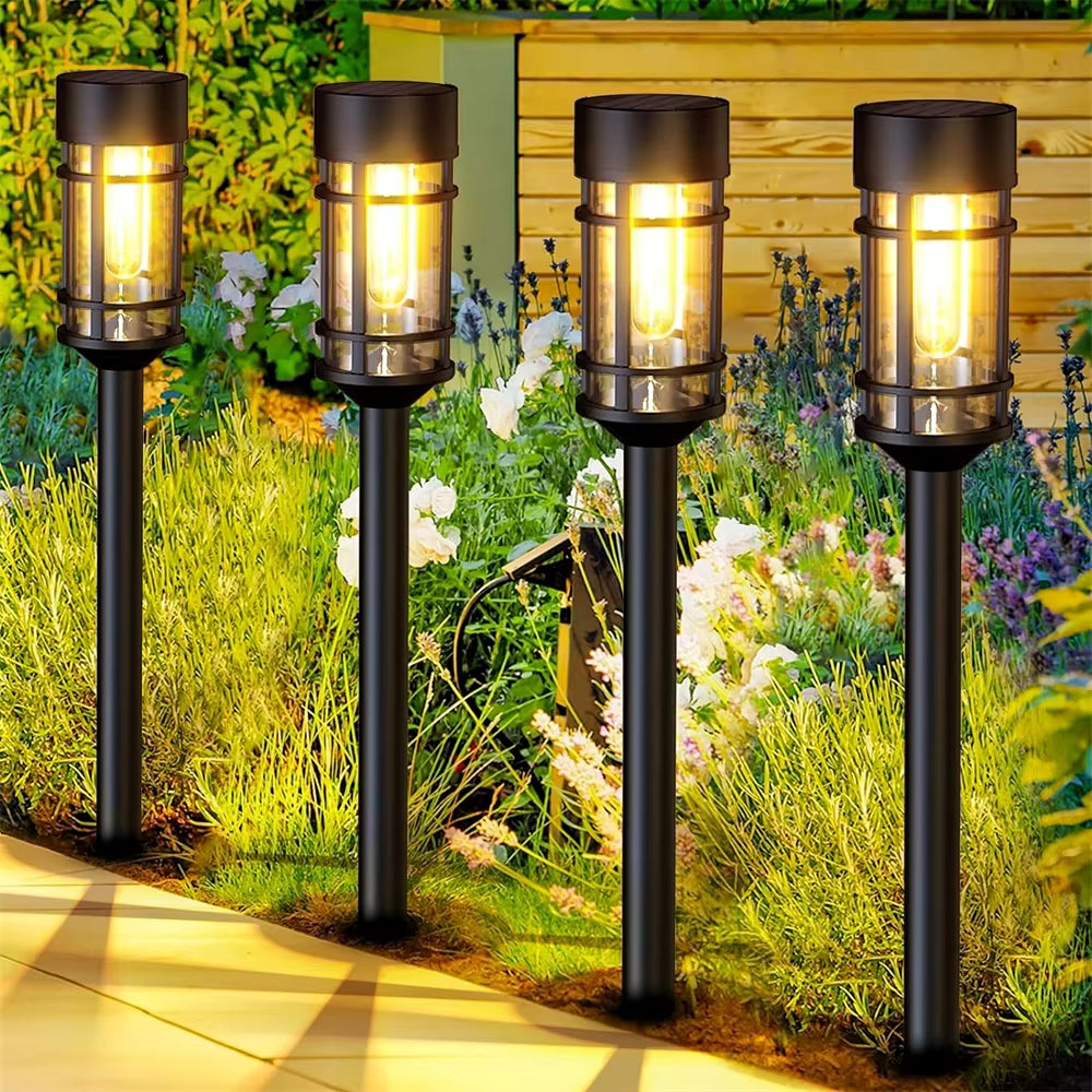 Four black outdoor solar post lights with clear glass shades are illuminated in a garden setting next to a paved walkway.