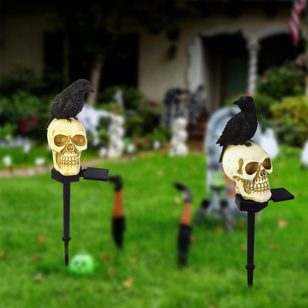 Two Halloween yard decorations featuring black raven figurines perched on cream-colored skulls mounted on black metal stakes, set in a grassy yard with a blurred house background.