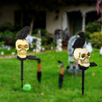 Two Halloween yard decorations featuring black raven figurines perched on cream-colored skulls mounted on black metal stakes, set in a grassy yard with a blurred house background.