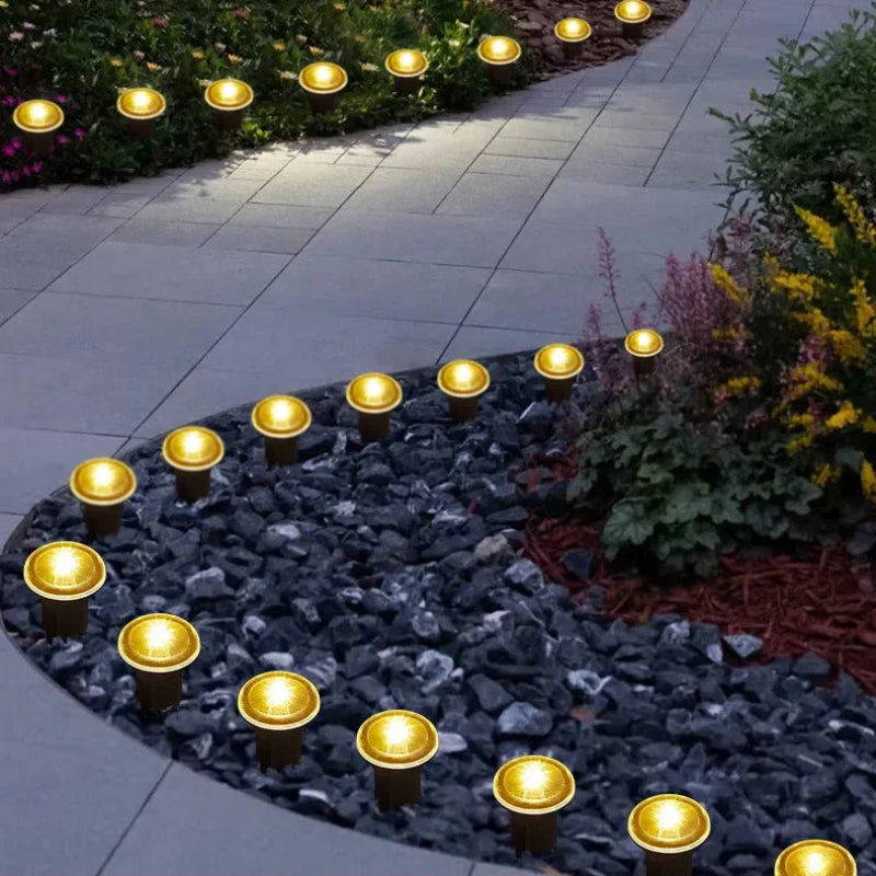 Solar landscape lights arranged along a paved walkway and garden beds at dusk, casting a warm yellow glow. The lights are mounted in curved rows over gray stone and mulch, with green plants and colorful flowers in the background.