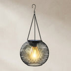 A spherical black metal mesh pendant light with a glowing warm Edison bulb inside, suspended by chains from a hook against a neutral beige background.