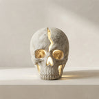 A skull-shaped decorative lamp with a textured gray concrete-like finish, featuring cutout openings that glow warm yellow from the internal light, set against a neutral light gray background.