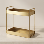 A two-tier gold metal rolling cart with integrated handles and open sides, shown on a light stone surface against a neutral beige background.