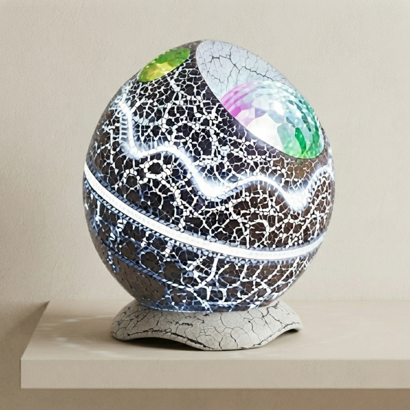 A decorative spherical lamp or orb with a cracked black and white ceramic-like surface, glowing white light bands, colorful mirrored facets near the top, and a textured light gray base, displayed on a beige surface against a beige wall.