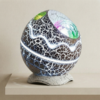 A decorative spherical lamp or orb with a cracked black and white ceramic-like surface, glowing white light bands, colorful mirrored facets near the top, and a textured light gray base, displayed on a beige surface against a beige wall.