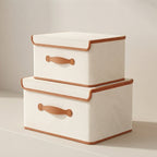 Two fabric storage boxes stacked on a light beige surface against a neutral cream background. The boxes are off-white with tan leather-like handles and trim.