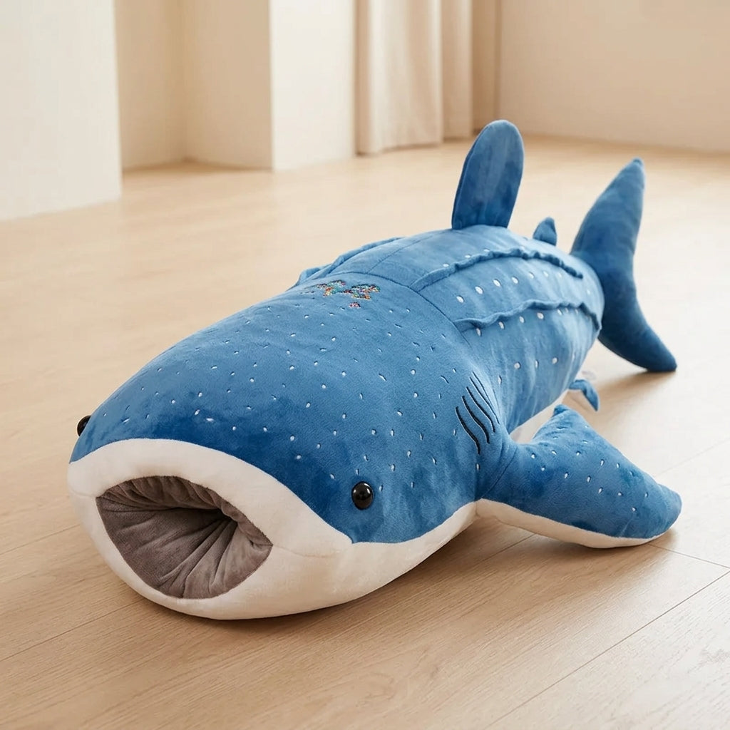 A plush shark-shaped floor cushion or pet bed in blue and white, photographed on a light wood floor in a softly lit room.