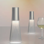 Two modern glass lamps on a reflective surface with a glass of white wine in the background.