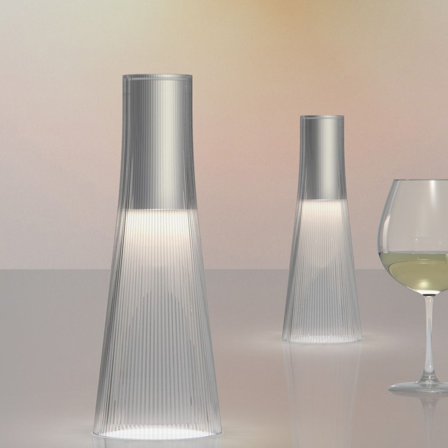 Two modern glass lamps on a reflective surface with a glass of white wine in the background.