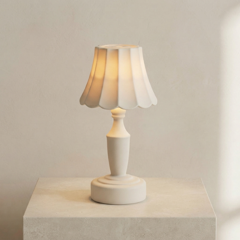 A small table lamp with a pleated white shade and a matching white pedestal base, shown lit against a soft beige wall on a light stone pedestal.
