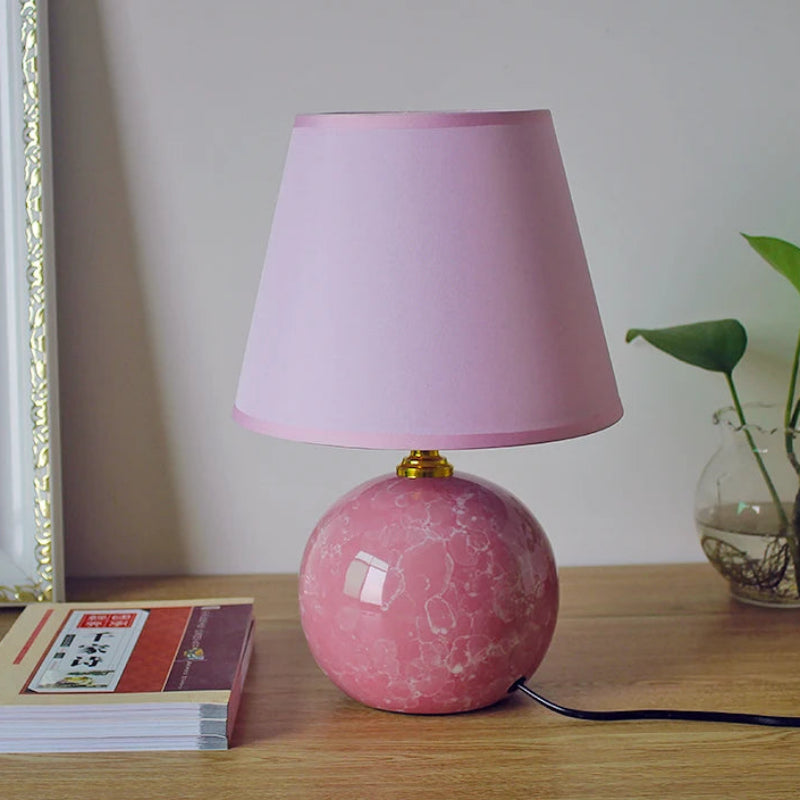 Pink marble table lamp with a pink shade on a wooden surface next to books and a plant.