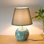 Table lamp with a green marble base and brown shade on a wooden surface, next to a plant.