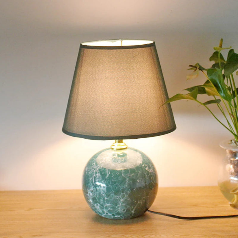 Table lamp with a green marble base and brown shade on a wooden surface, next to a plant.
