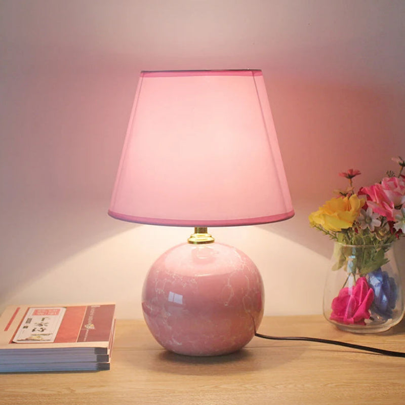 Pink table lamp on a wooden surface with a vase of flowers in the background