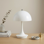 A modern white mushroom-shaped table lamp sits on a light wood tabletop against a warm beige wall. Decorative props include a small vase with dried stems, stacked books, a gray tray with stones, and a gold cylindrical object.